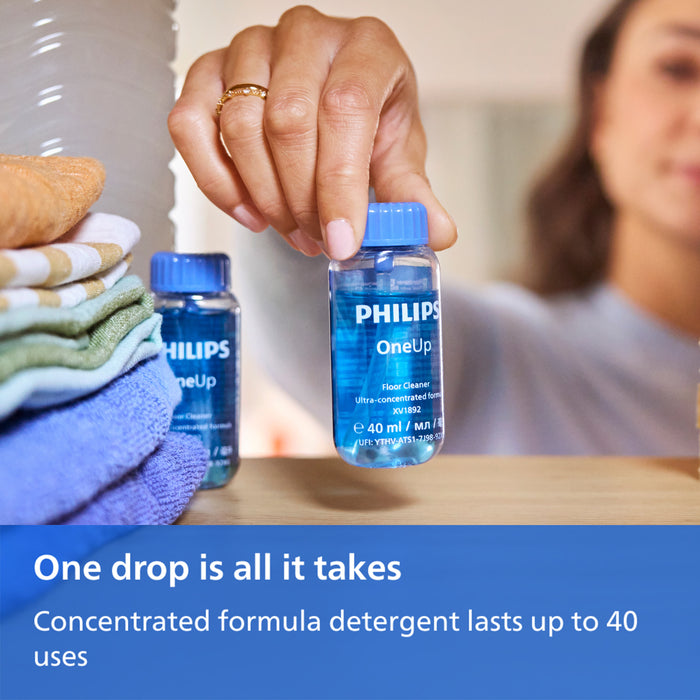 A person's hand holds a small blue Philips OneUp floor cleaner bottle over a wooden surface, with another bottle and folded towels in the background. A blue text banner at the bottom reads, 'One drop is all it takes.